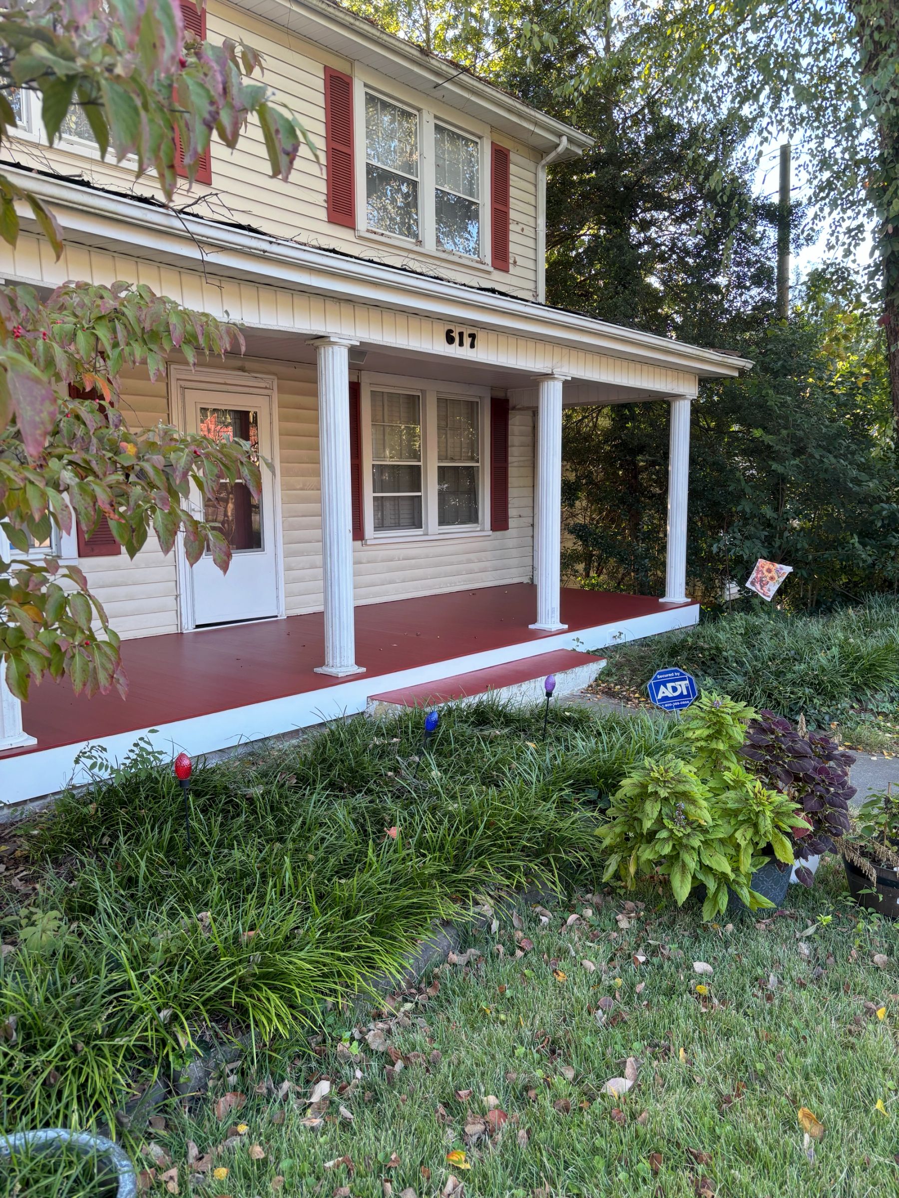 A classic front porch with white columns and a red-painted floor complements the home’s cream siding, red shutters and leafy surroundings.